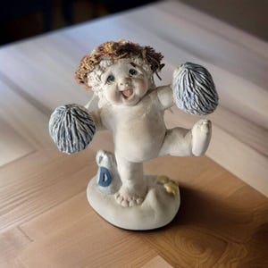 May include: A white cherub figurine with brown hair, holding blue pom-poms and standing on a base with a blue letter 'D'.
