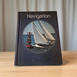 Op de afbeelding: Een vintage boek met de titel "Navigation" met een marineblauwe omslag. De omslag toont een cirkelvormige afbeelding van een zeilboot met witte zeilen en een rode boei. Het boek staat rechtop op een lichtbruin oppervlak.