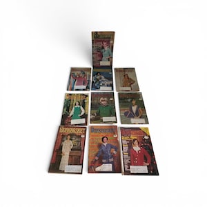 May include: A collection of ten vintage The Workbasket magazines arranged in a pyramid formation. Each magazine features a colorful cover with various craft and fashion photography, including titles and text printed on the front covers.