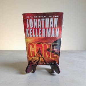 May include: A hardcover book titled "GONE" by Jonathan Kellerman, displayed on a small, dark red wooden stand. The book cover features a landscape with a road and the author's name in large, silver letters. The stand has a simple, functional design.