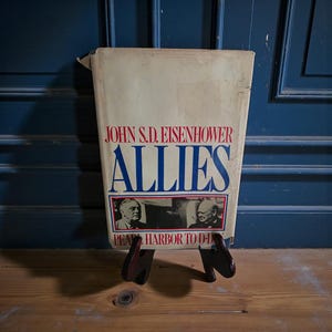 May include: A vintage book titled "ALLIES" by John S.D. Eisenhower, displayed on a wooden stand. The book cover features red and blue text, with images of historical figures. The subtitle reads "PEARL HARBOR TO D-DAY".