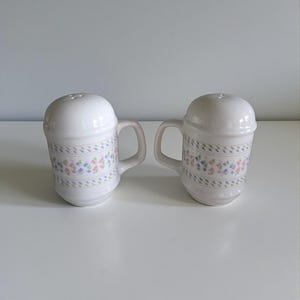Puede incluir: Dos saleros y pimenteros de cerámica blanca con asas. Cada salero tiene una tapa abovedada y está decorado con un patrón geométrico pastel en rosa, azul y verde. Los saleros están diseñados para parecerse a pequeñas tazas.