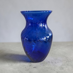 May include: A vibrant cobalt blue glass vase with a smooth, translucent finish. The vessel features a wide, flared rim that tapers into a rounded, bulbous body, creating a classic silhouette. The glass surface reflects light, highlighting its deep color.