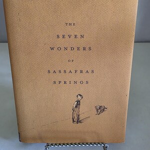 Może przedstawiać: Zabytkowa książka zatytułowana "The Seven Wonders of Sassafras Springs" z beżową okładką. Okładka zawiera czarny tekst i małą ilustrację chłopca i psa. Książka jest prezentowana na metalowym stojaku.