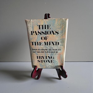 May include: A vintage book titled "The Passions of the Mind" by Irving Stone, displayed on a small, dark red wooden easel. The book cover features a marbled design in shades of blue, beige, and peach. The text includes "A Biographical Novel of Sigmund Freud."
