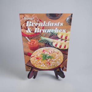 Könnte beinhalten: Ein Vintage-Kochbuch mit dem Titel "Sunset Breakfasts & Brunches" wird auf einem Holzständer präsentiert. Das Cover zeigt Bilder von Frühstücks- und Brunchgerichten, darunter Gebäck, einen Auflauf und Obst.
