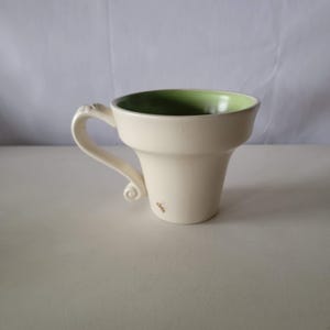 May include: Cream-colored ceramic mug with a flared shape and a green interior. The handle has a decorative swirl at the base. A small, red floral design is on the side of the mug. The mug is designed for hot beverages.