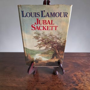 May include: A vintage copy of "Jubal Sackett" by Louis L'Amour, displayed on a brown stand. The book cover features a landscape painting with a river, trees, and the title in blue and red lettering. The book is slightly worn.