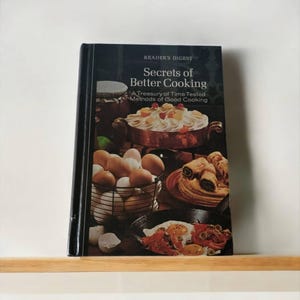 May include: A black cookbook with a white title that reads "Secrets of Better Cooking: A Treasury of Time Tested Methods of Good Cooking". The cover features a photo of a basket of eggs, a bowl of pastry, and a casserole dish with a creamy topping.