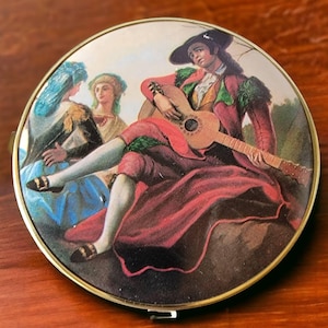 May include: Vintage round compact mirror with a painted scene of a man playing a guitar, dressed in red and black clothing, with two women in the background. The compact has a gold-colored metal frame.
