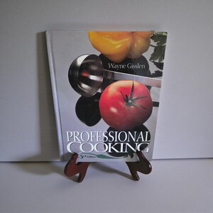 May include: A cookbook titled "Professional Cooking" by Wayne Gisslen, 3rd Edition, is displayed on a wooden stand. The cover features a red tomato, yellow bell pepper, and a stainless steel scoop. The book is propped up on a brown wooden stand.