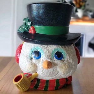 Puede incluir: Una figura de muñeco de nieve blanco con un sombrero de copa negro, acebo verde, orejeras rojas y una pipa amarilla. El muñeco de nieve tiene ojos azules, una bufanda a rayas rojas y verdes y una sonrisa pintada.