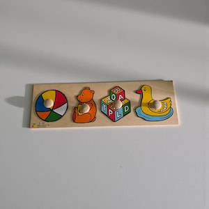 May include: A rectangular wooden peg puzzle board featuring four colorful illustrations: a multi-colored ball, an orange bear, a cube with the letters B, O, A, T, P, L, D, and a yellow duck on blue water. Each piece has a small round wooden knob.