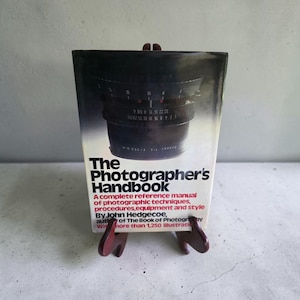 May include: A hardcover book titled The Photographer's Handbook by John Hedgecoe, featuring a black and white photograph of a camera lens on the cover. The text includes a subtitle about photographic techniques, equipment, and style.