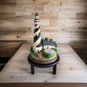 Könnte beinhalten: Dekorative Leuchtturm-Figur mit einem schwarz-weißen Spiraldesign, einem kleinen Haus und einer kreisförmigen Schüssel. Der Leuchtturm hat eine rote Spitze und steht auf einem Sockel mit Grün. Die Figur steht auf einem kleinen schwarzen Ständer.