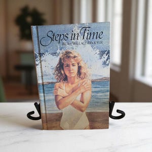 Puede incluir: Un libro antiguo titulado "Steps in Time" de Ruth Wallace Brodeur. La portada presenta a una mujer en traje de baño blanco con los brazos cruzados, ambientada en una escena de playa con olas rompiendo. El libro se muestra en un soporte de metal negro.