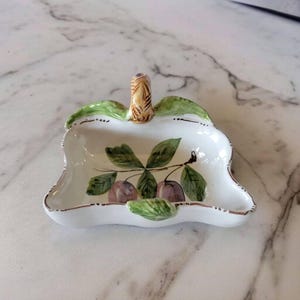 Könnte beinhalten: Eine weiße Keramikschale mit einem Griff in Form eines Zweigs mit grünen Blättern und lila Pflaumen. Die Schale hat einen gewellten Rand und einen goldenen Rand.