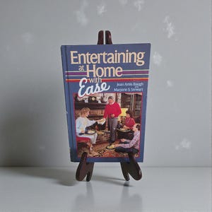 Puede incluir: Un libro titulado "Entertaining at Home with Ease" se muestra en un caballete de madera. La portada del libro es azul con texto blanco y rojo y una foto de gente cenando. El caballete es de color marrón oscuro.