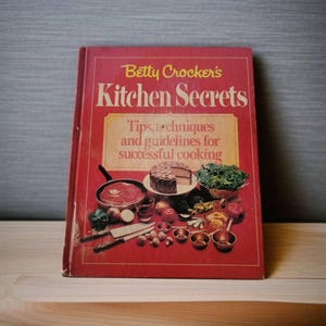 May include: A red cookbook titled "Betty Crocker's Kitchen Secrets: Tips, techniques and guidelines for successful cooking" with a photo of a cake, vegetables, and a skillet on the cover.