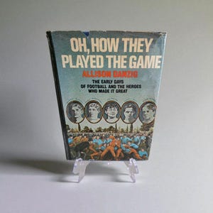 Op de afbeelding: Een vintage boek getiteld "Oh, How They Played the Game" van Allison Danzig. De omslag heeft een blauw en wit ontwerp met portretten van voetballers en een illustratie van een voetbalwedstrijd. Het boek wordt gepresenteerd op een transparante plastic standaard.