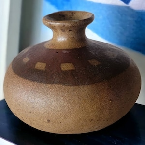 May include: A brown ceramic vase with a wide, rounded body and a narrow neck. The vase has a brown and tan glaze with a band of brown squares around the middle.