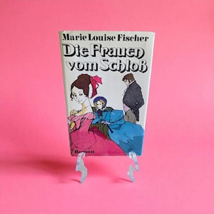 May include: A vintage book titled "Die Frauen vom Schloß" with a cover illustration of three figures in period clothing. The cover features a woman in a pink dress, a woman in a blue dress, and a man in a black coat. The book is propped up on a clear stand against a pink background.
