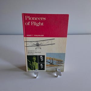 May include: A vintage book titled "Pioneers of Flight" with a red cover and a cream-colored spine. The cover features an illustration of an early airplane and the text "Compliments of Disabled American Veterans."