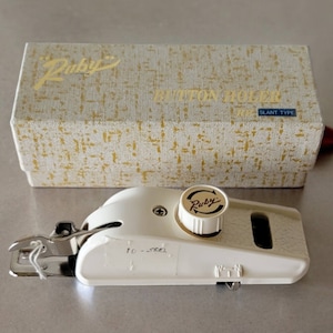 May include: White buttonhole maker with a white knob that says "Ruby" in brown lettering. The buttonhole maker is in a white box with the words "Ruby Button Holer Slant Type" printed on the top.