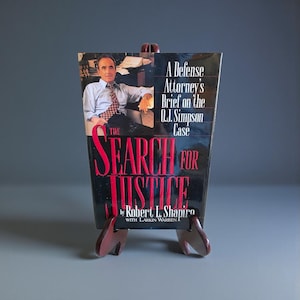 May include: A book titled "The Search for Justice" by Robert L. Shapiro, displayed on a small wooden easel. The book cover features a photo of a man and text that reads "A Defense Attorney's Brief on the O.J. Simpson Case."