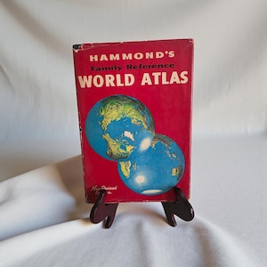 May include: A vintage "HAMMOND'S Family Reference WORLD ATLAS" book with a red cover and two globes. The book is displayed on a small wooden stand. The atlas is a New Revised Edition.