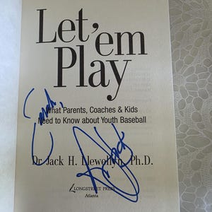 Op de afbeelding: Een witte boektitelpagina met zwarte tekst die Let'em Play, What Parents, Coaches & Kids Need to Know about Youth Baseball en Dr. Jack H. Llewellyn Ph.D. vermeldt. De pagina bevat twee duidelijke handtekeningen in blauwe inkt.