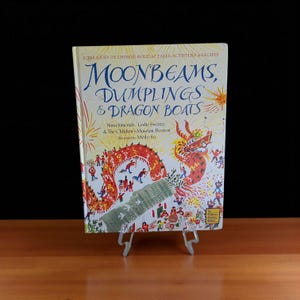 May include: A children's book titled "Moonbeams, Dumplings & Dragon Boats" on a clear stand. The cover features a red dragon illustration with people and fireworks. The book is a treasury of Chinese holiday tales, activities, and recipes.