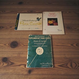 May include: Three vintage piano course books are displayed on a wooden surface. The books include "Alfred d'Auberge Piano Course", "Musicianship for the Older Beginner", and "John Thompson's Supplementary Piano Course". The books have aged covers and text.
