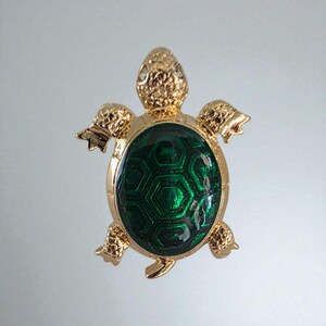May include: A gold-toned turtle brooch with a textured shell and limbs. The turtle's shell is a deep green with a geometric pattern. The brooch is a decorative accessory.