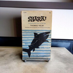 May include: Vintage book titled "SHARK! Unpredictable Killer of the Sea" by Thomas Helm. The cover features a black shark illustration over blue and white horizontal stripes. The book is displayed on a metal stand.