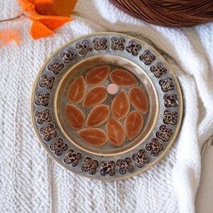 May include: A round, decorative dish with a gold rim and a brown and black patterned border. The center of the dish features a design of brown leaves.