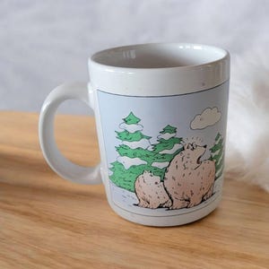 May include: White ceramic mug featuring a cartoon illustration of two bears in a snowy forest with green trees. The mug has a curved handle and a light blue background.