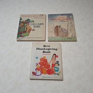 May include: Three vintage children's books are displayed. The covers feature illustrations and titles: "Log-Cabin Home," "A book about Christopher Columbus," and "Best Thanksgiving Book." The books are on a light-colored surface.