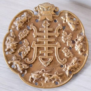 May include: A detailed, bronze-colored, circular pendant with intricate relief carvings. The design includes a central Chinese character, surrounded by mythical creatures and cloud motifs. The pendant has a decorative top element and small circular holes.