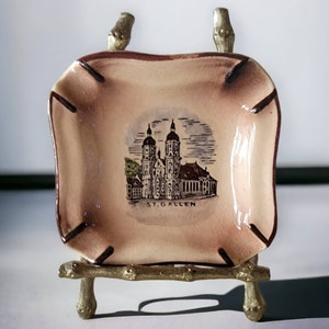 May include: A small, square, ceramic ashtray with a brown and white design of a church building. The ashtray is on a small, gold-colored stand. The text "ST. GALLEN" is printed on the ashtray.