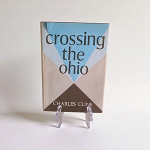 May include: A vintage book titled "Crossing the Ohio" by Charles Cline. The cover has a geometric design with a blue triangle, beige and brown shapes, and the title in a dark font. The book is displayed on a clear plastic stand, ideal for a collector.