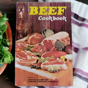 以下が含まれることがあります： 茶色の表紙に「BEEF Cookbook」というタイトルが黄色い文字で書かれたビンテージの料理本。表紙には、さまざまな牛肉の切り身、野菜、ハーブの写真が掲載されています。