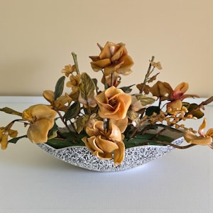 May include: A decorative floral arrangement featuring amber-colored sculpted flowers and green leaves set in a low, boat-shaped planter with a speckled black and white finish. The arrangement includes multiple rose-like blooms and small sprigs.