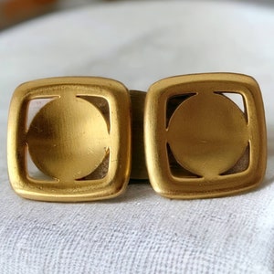 May include: A pair of gold-toned earrings with a square shape and a circular cutout in the center. The earrings have a brushed finish.