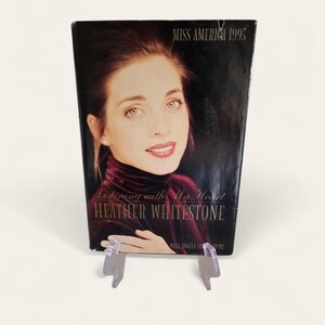 Puede incluir: Un libro titulado "Listening with My Heart" de Heather Whitestone, con "Miss America 1995" impreso en la parte superior. La portada presenta un retrato de una mujer con cabello oscuro y una blusa burdeos.