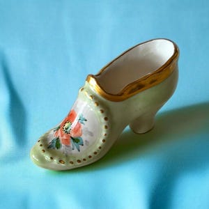 May include: A small, ceramic shoe figurine in a light green hue with gold trim. The shoe features a painted floral design in shades of orange and green on the front. The heel is curved, and the shoe is approximately 4 inches long.