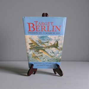 May include: A book titled "Target Berlin" with a light blue cover and a depiction of WWII-era bomber planes. The book is propped up on a small wooden easel. The text on the cover reads "Mission 250: 6 March 1944".
