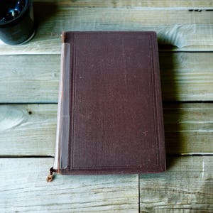 Puede incluir: Un libro encuadernado en cuero marrón con un lomo que tiene una etiqueta blanca. El libro está sobre una superficie de madera.