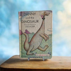 Puede incluir: Un libro infantil vintage titulado "Danny and the Dinosaur" de Syd Hoff. La portada presenta una ilustración de un dinosaurio amigable con un niño en su cuello. El libro se muestra en un pequeño soporte de metal.