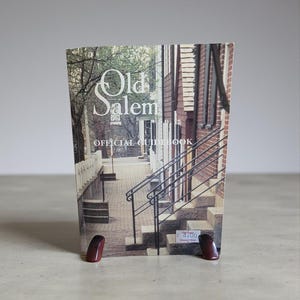 May include: An "Old Salem Official Guidebook" is propped up by two small, dark red bookends. The cover displays a photograph of a brick building with steps and a railing. The title is in a decorative font.